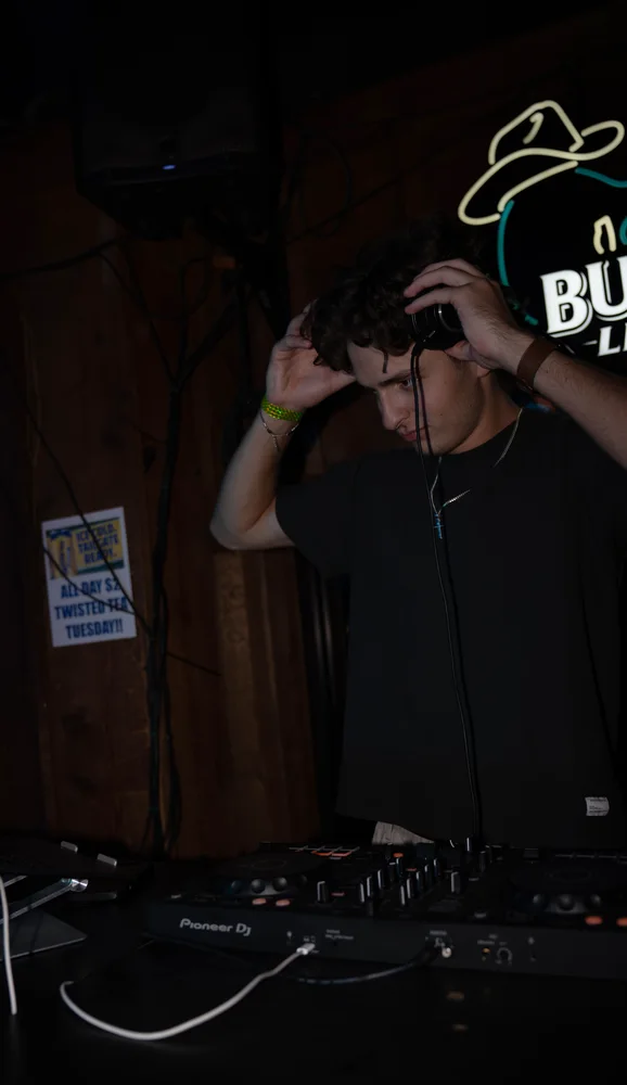 Lane Watt raising a hand towards his headphones at the DJ booth